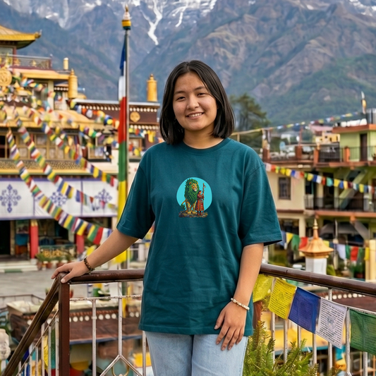 Person wearing a teal t-shirt with a graphic design in front of a traditional building and mountainous background.