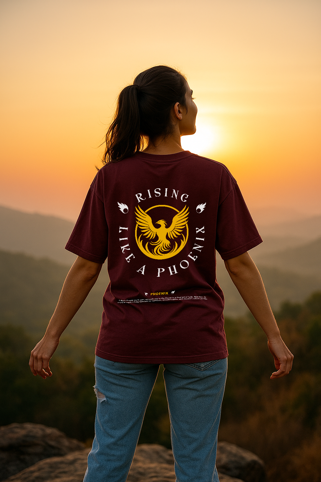 Person wearing a maroon t-shirt with a phoenix design, standing on a mountain at sunset.