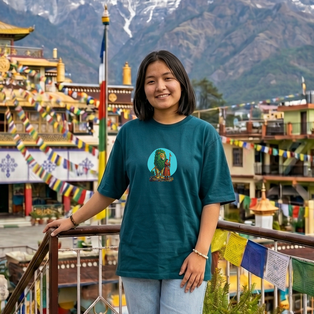 Person wearing a teal t-shirt with a graphic design in front of a traditional building and mountainous background.