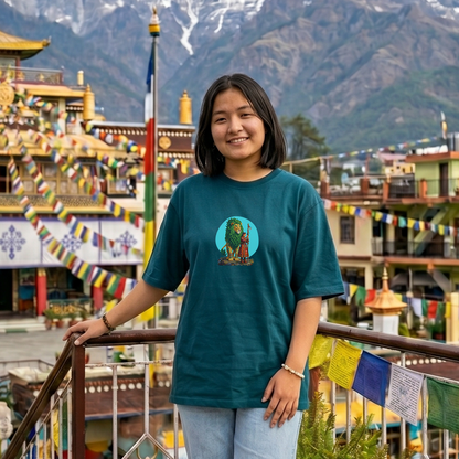 Person wearing a teal t-shirt with a graphic design in front of a traditional building and mountainous background.